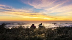 Two people sitting on the beach at sunset, overlooking the ocean.