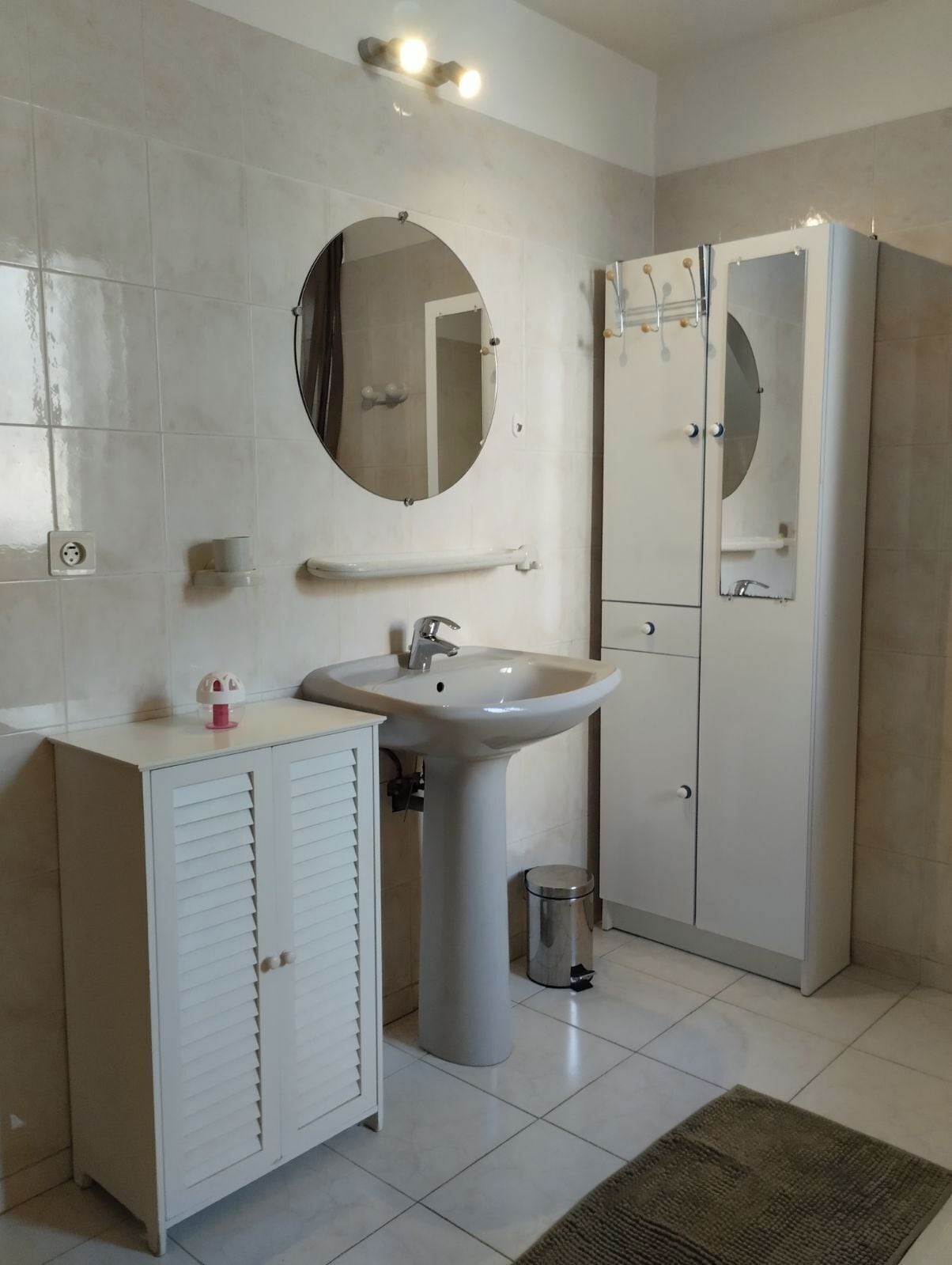 A bathroom with a white sink, mirror, and cabinet, a trash bin, and a shower area.