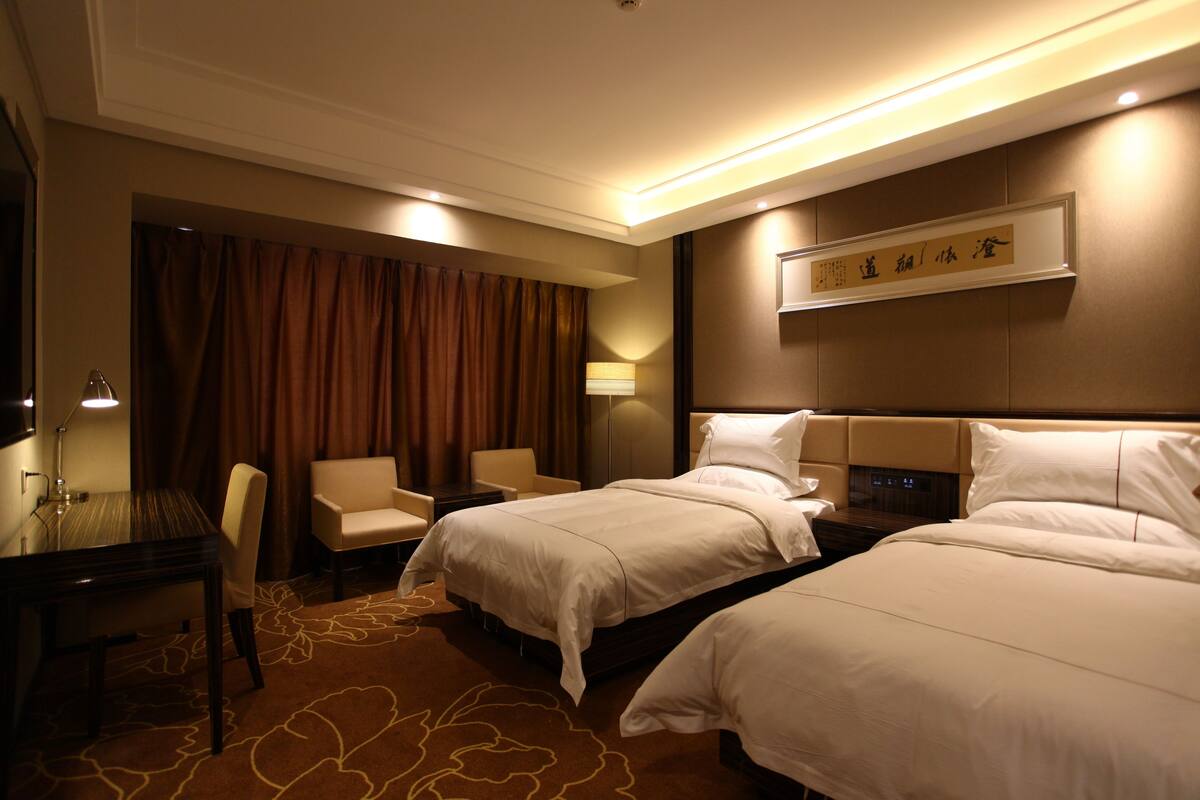 Ein Hotelzimmer mit zwei Betten, einem Schreibtisch, einem Stuhl, einer Lampe und einem an der Wand befestigten Schild.