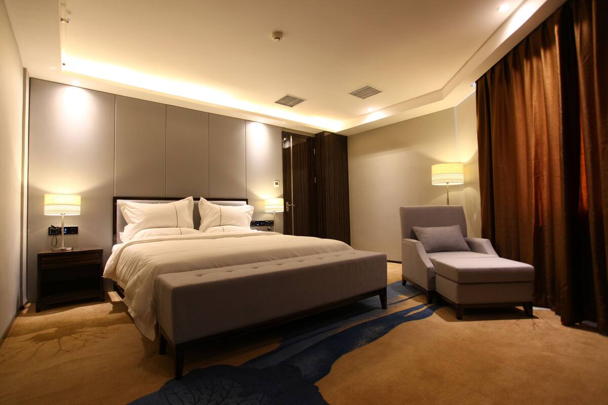 Ein modernes Hotelzimmer mit einem großen Bett, einem Nachttisch mit Lampe, einem Sessel und Teppichboden.