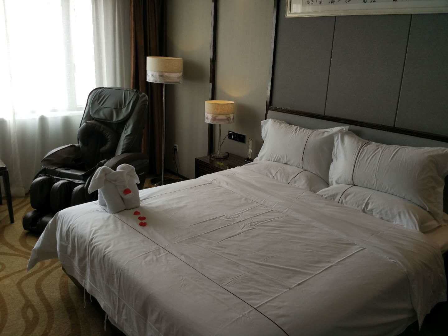 A hotel room with a large bed, a massage chair, a lamp, and a small table.