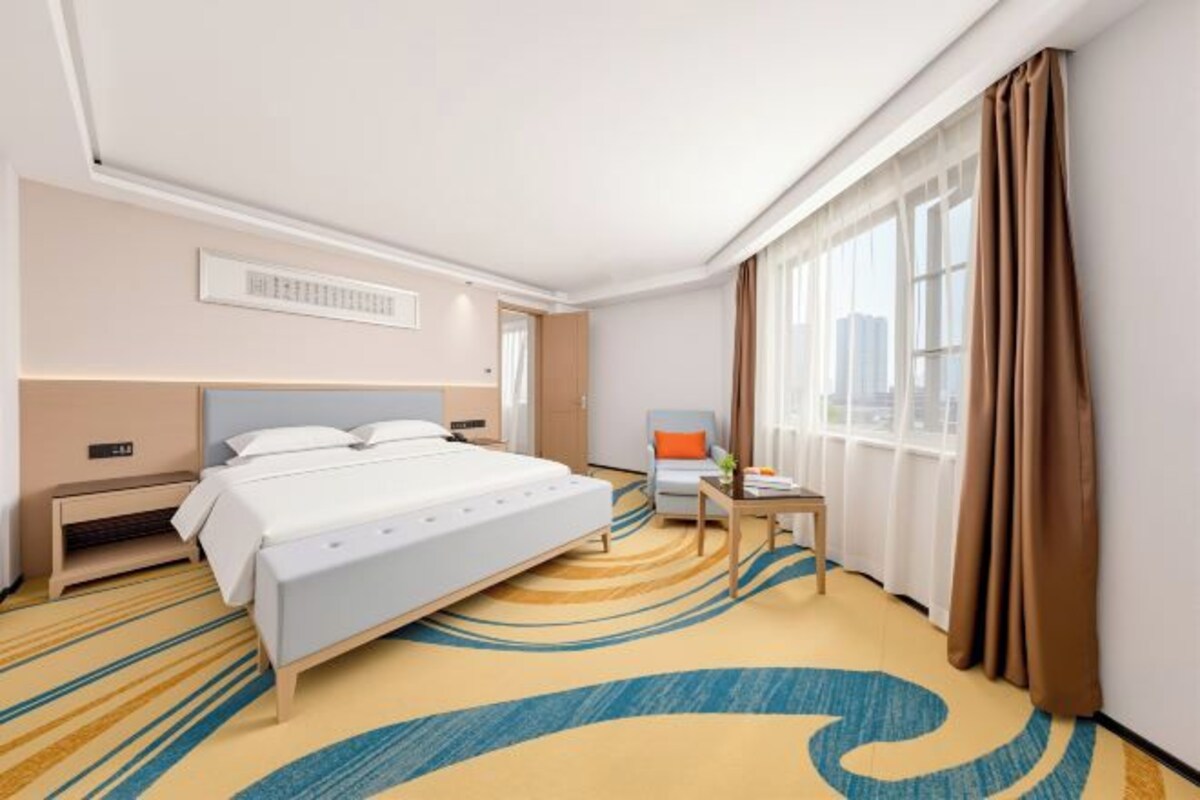 A hotel room with a bed, a desk, a chair, a window with curtains, and a patterned carpet.