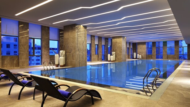 An indoor swimming pool with modern lighting, lounge chairs, and large windows offering city views. - Wanda Realm Dongying (Dongying)