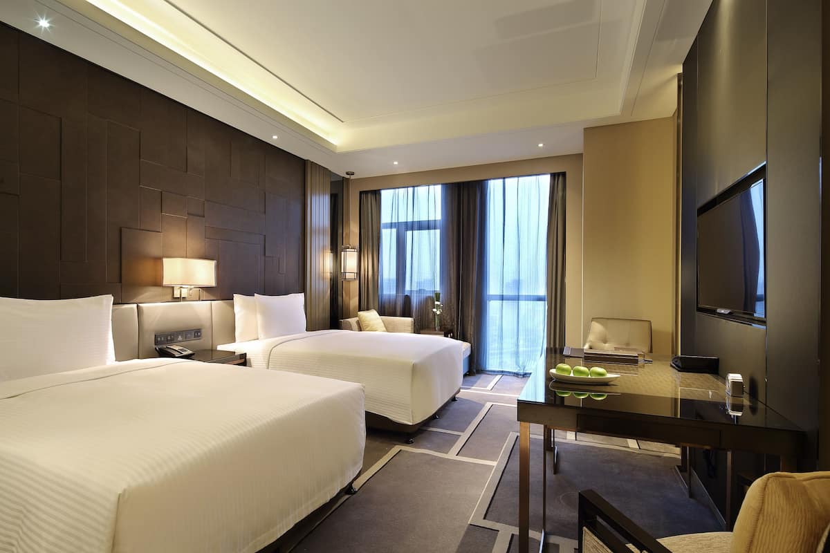 A hotel room with two beds, a desk, a TV, and a large window with curtains.