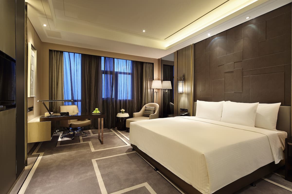 A modern hotel room with a large bed, a desk with a chair, a small table with a lamp, and a view of the city through large windows.