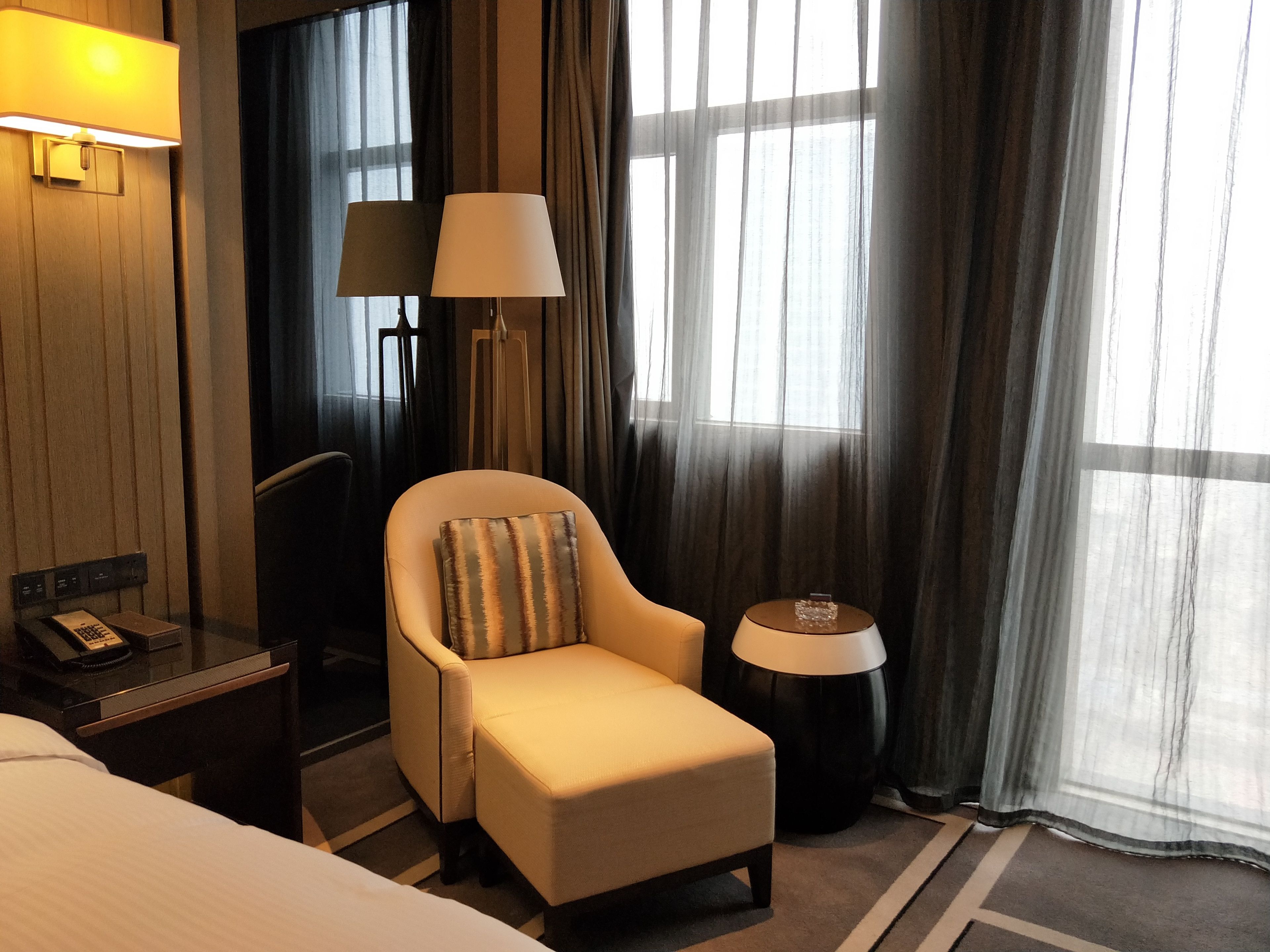 Une chambre d’hôtel avec un lit, une chaise avec un coussin, une table de chevet avec un téléphone, une lampe et une fenêtre avec des rideaux.