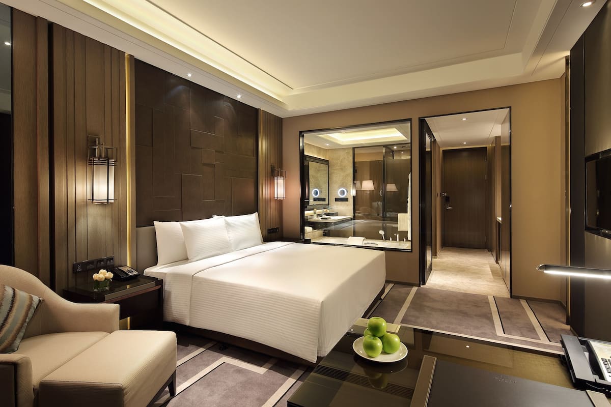 A modern hotel room with a large bed, a sitting area, a desk with a lamp, and a mirror.