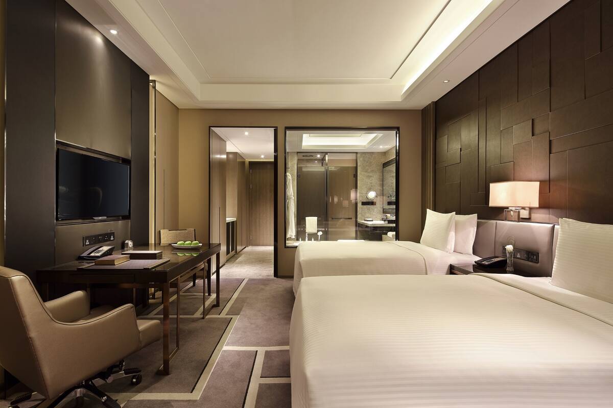 A hotel room with two beds, a desk with a computer, a TV, and a bathroom with a mirror and sink.