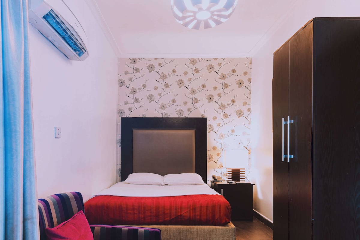 A bedroom with a bed, a nightstand, a wardrobe, and a window with floral wallpaper.