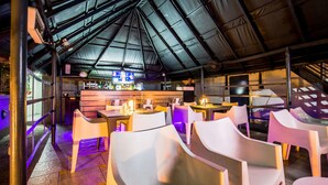 Un comedor interior con sillas y mesas blancas, una barra con botellas y un techo alto con vigas de metal.