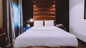Un cuarto de hotel con una cama grande, una mesita de noche con lámpara, una silla roja y vistas de la ciudad a través de la ventana.