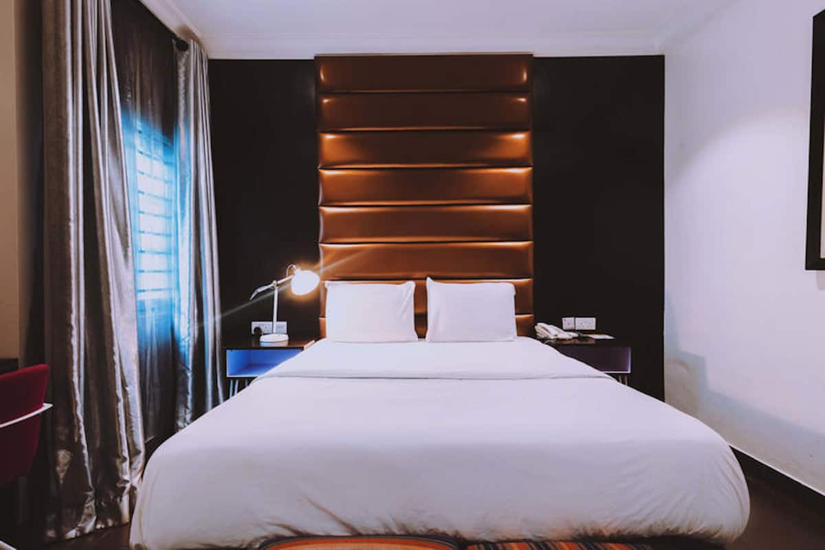 A hotel room with a large bed, a nightstand with a lamp, a red chair, and a view of the city through the window.