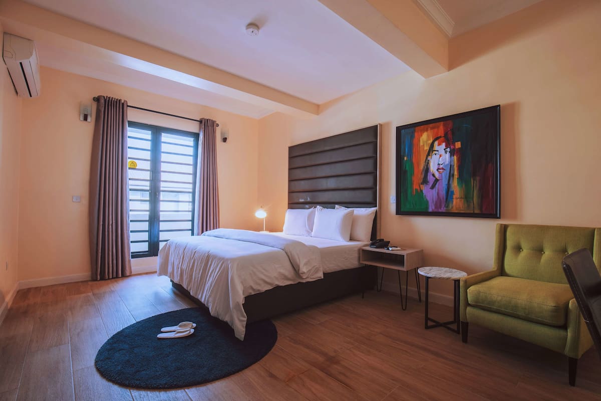 A hotel room with a bed, a nightstand, a green armchair, a small table, and a painting on the wall.