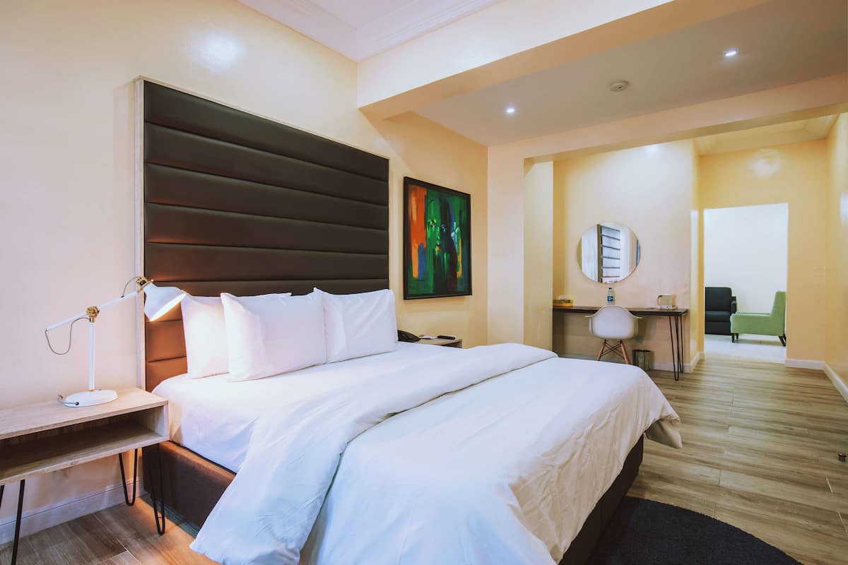A hotel room with a large bed, bedside table with a lamp, a desk with a chair, and a painting on the wall.