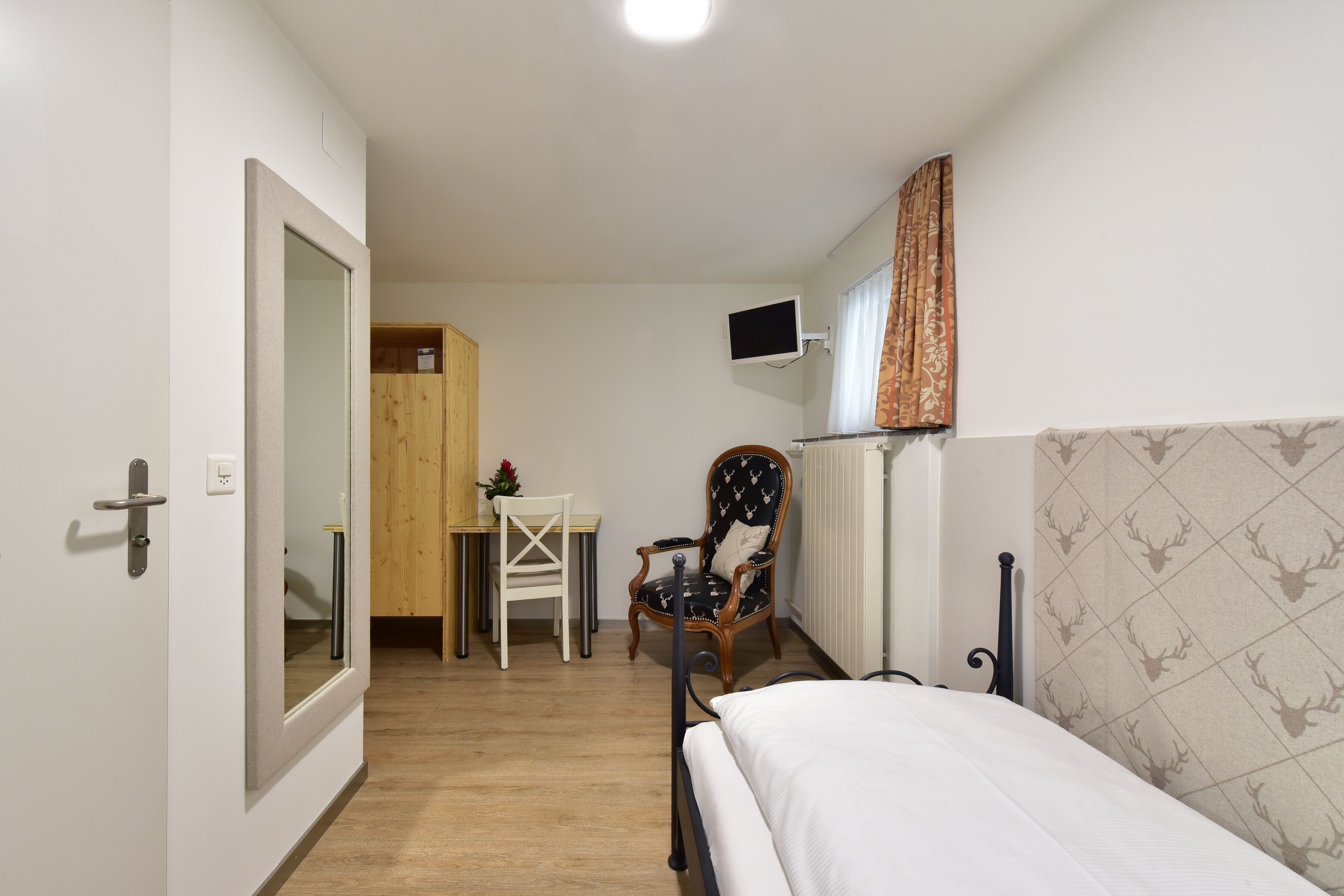 A compact room with a bed, a wooden chair, a small table, and a television mounted on the wall.