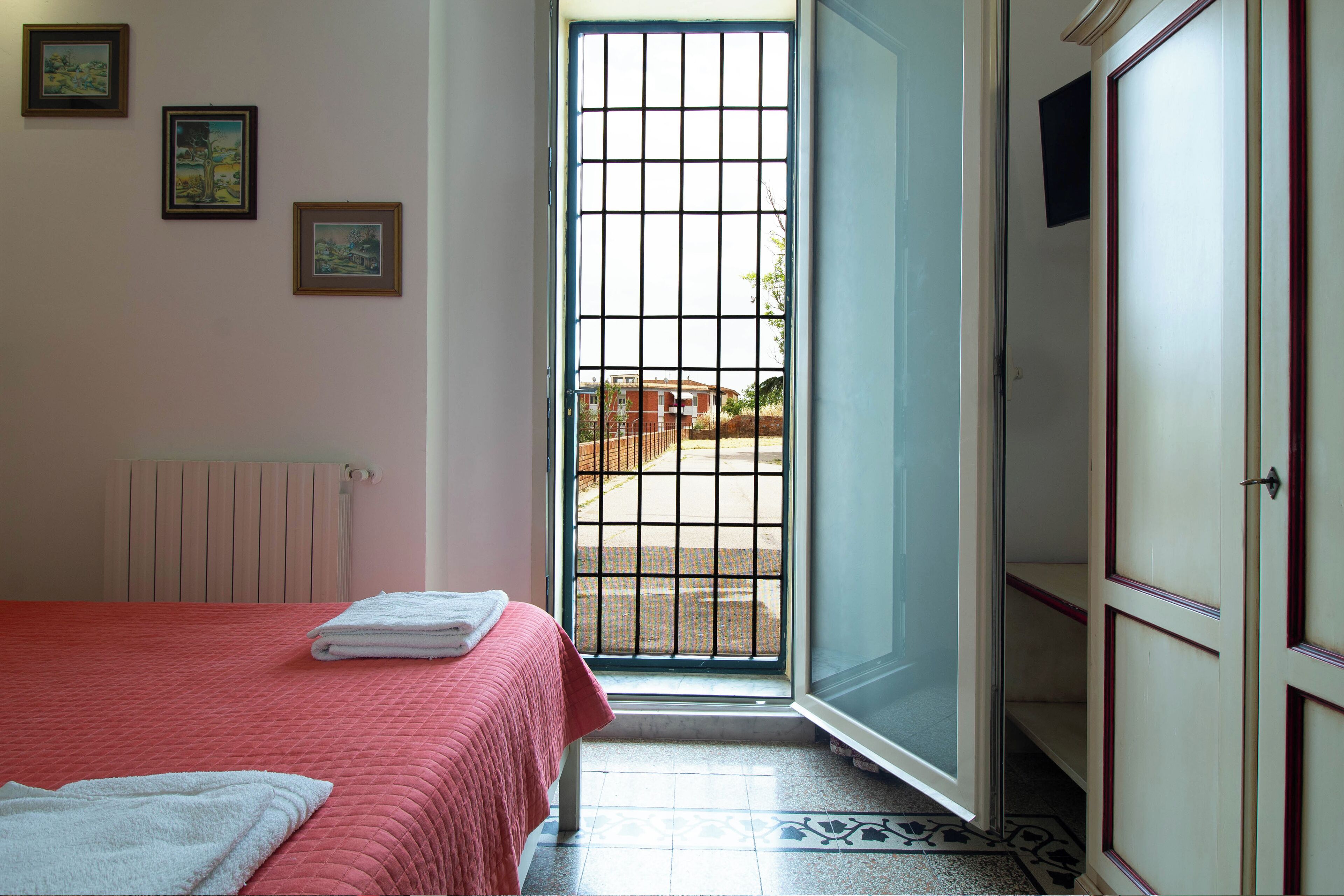 Una habitación de hotel con una cama, un radiador, una puerta con vistas y suelo de baldosas.