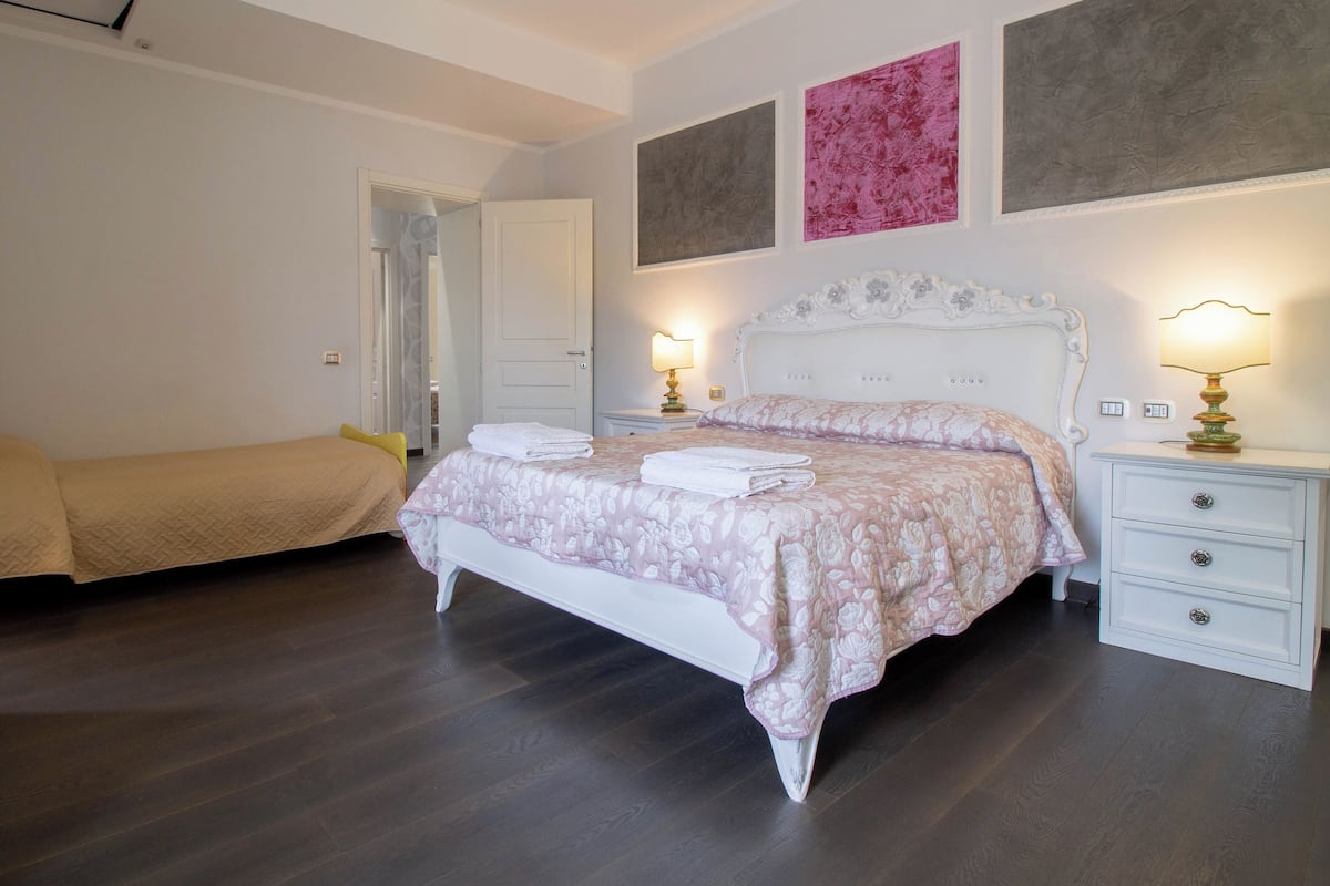 A bedroom with a large bed, two bedside tables, a nightstand, and a door leading to another room.