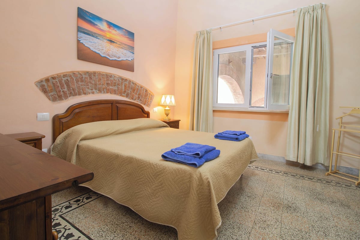 A bedroom with a bed, a nightstand, a lamp, a window with curtains, and a painting on the wall.