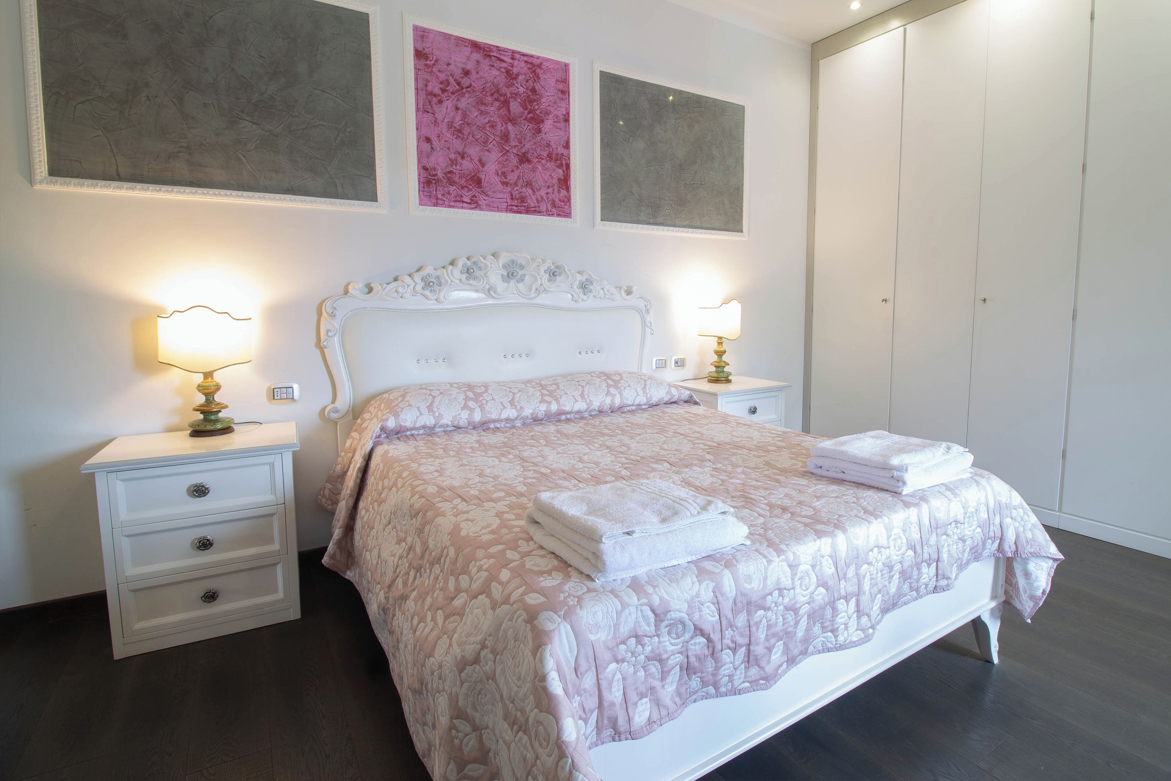 Un dormitorio con cama blanca, dos mesitas de noche con lámparas, un armario grande y dos cuadros en la pared.