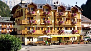 A multi-story hotel with balconies adorned with flower baskets, a restaurant area with outdoor seating, and a mountain backdrop.