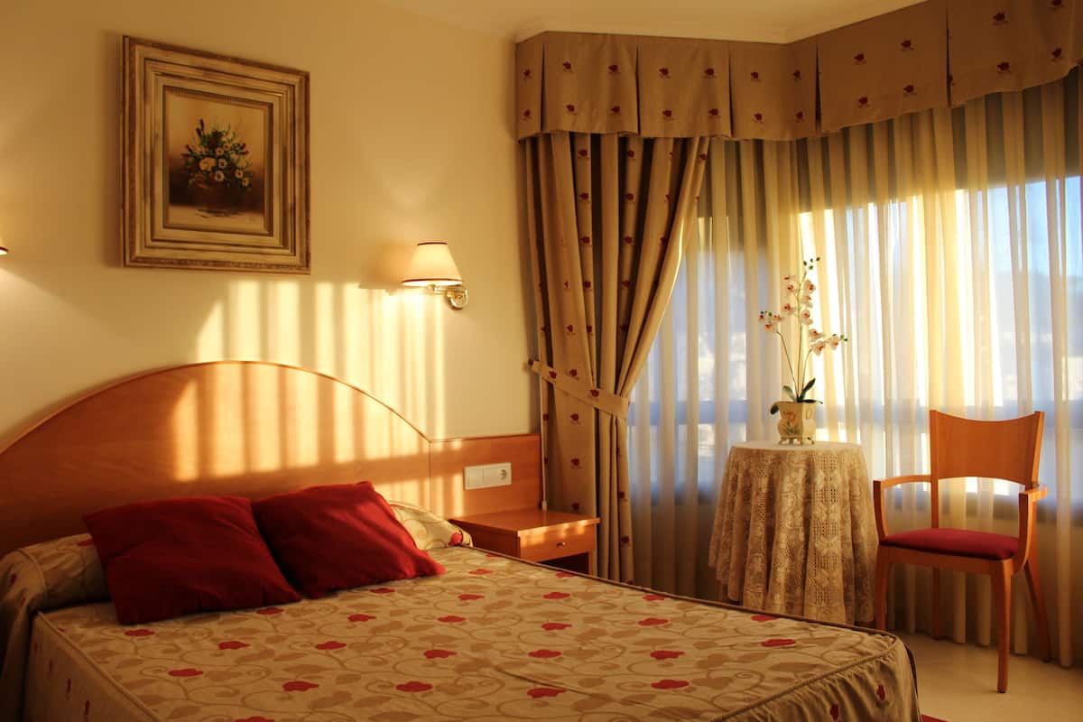 Une chambre d’hôtel avec un lit, une chaise, une table avec un vase et des rideaux.