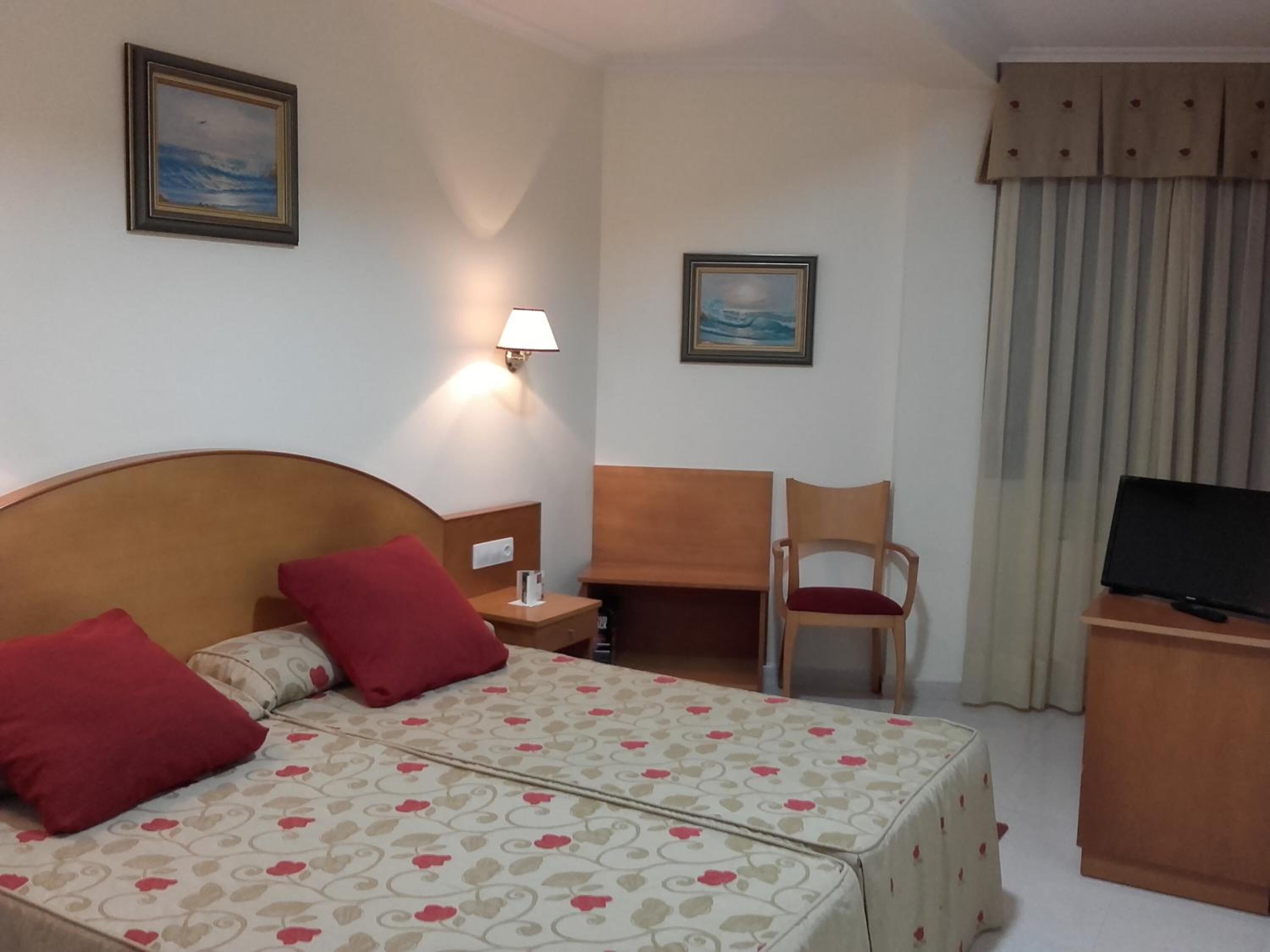 A hotel room with a bed, a desk, a TV, and two paintings on the wall.