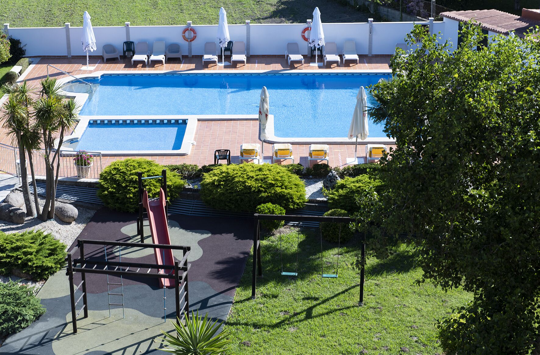 A swimming pool with lounge chairs and umbrellas, surrounded by greenery and a playground.