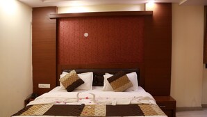 A neatly made bed with patterned bedding and two decorative pillows, a wooden headboard, and a bedside table with a power outlet. - Hotel Shivam International (New Delhi)