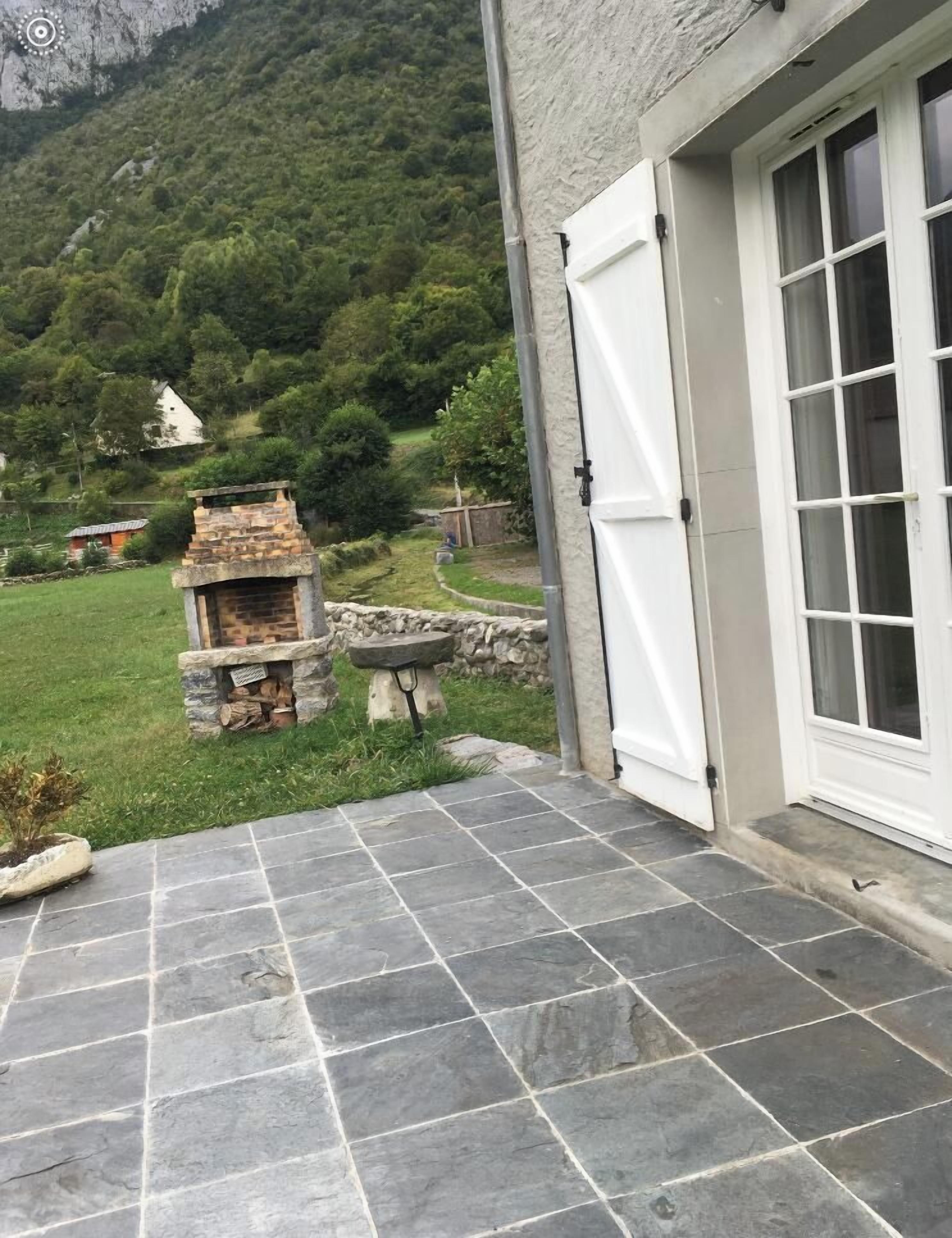Un patio de piedra con una chimenea de piedra, una puerta blanca y vistas a un paisaje verde con árboles y una montaña.
