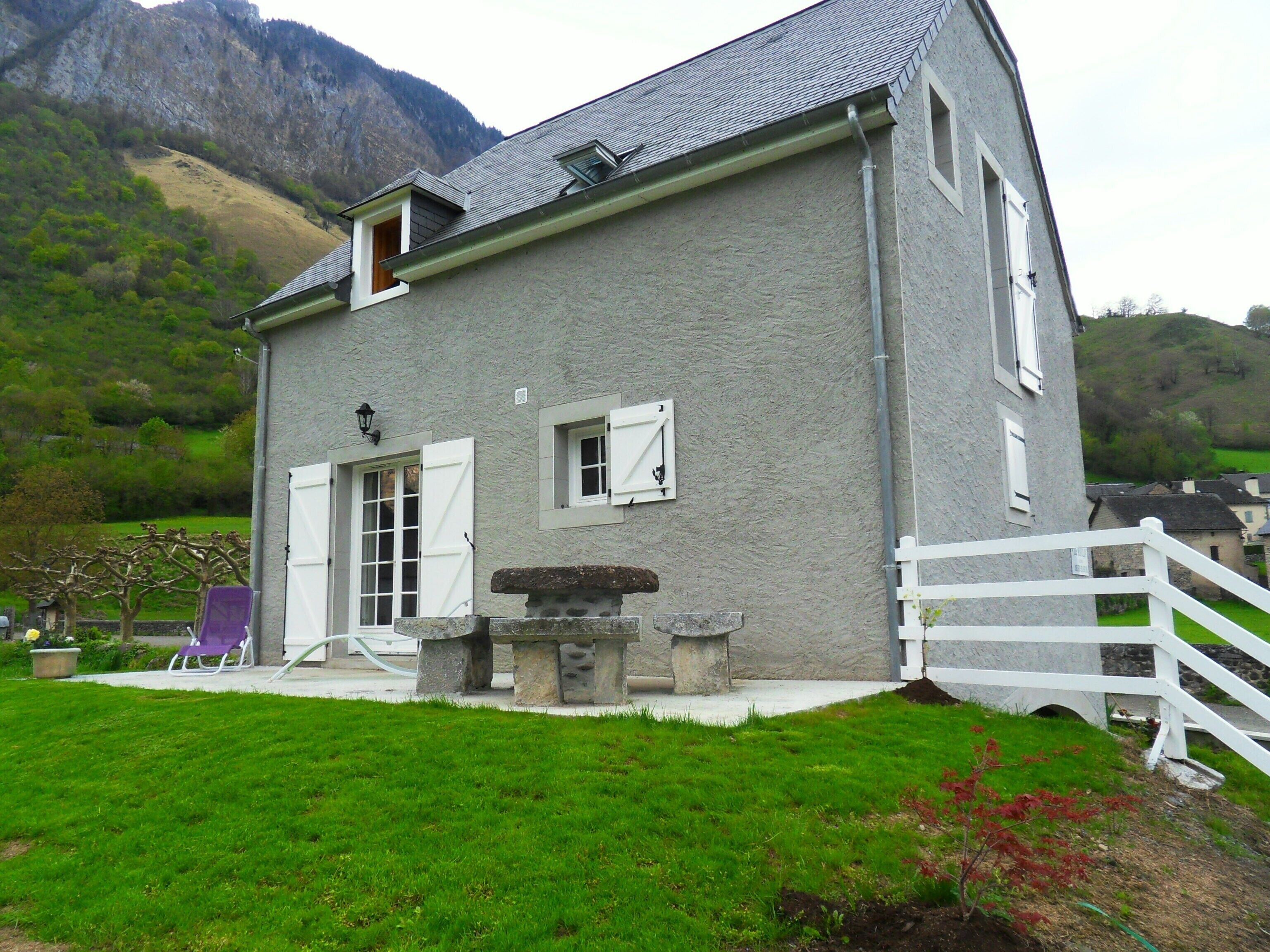 Une maison à deux étages avec un banc en pierre, une clôture blanche et une cour couverte d’herbe.