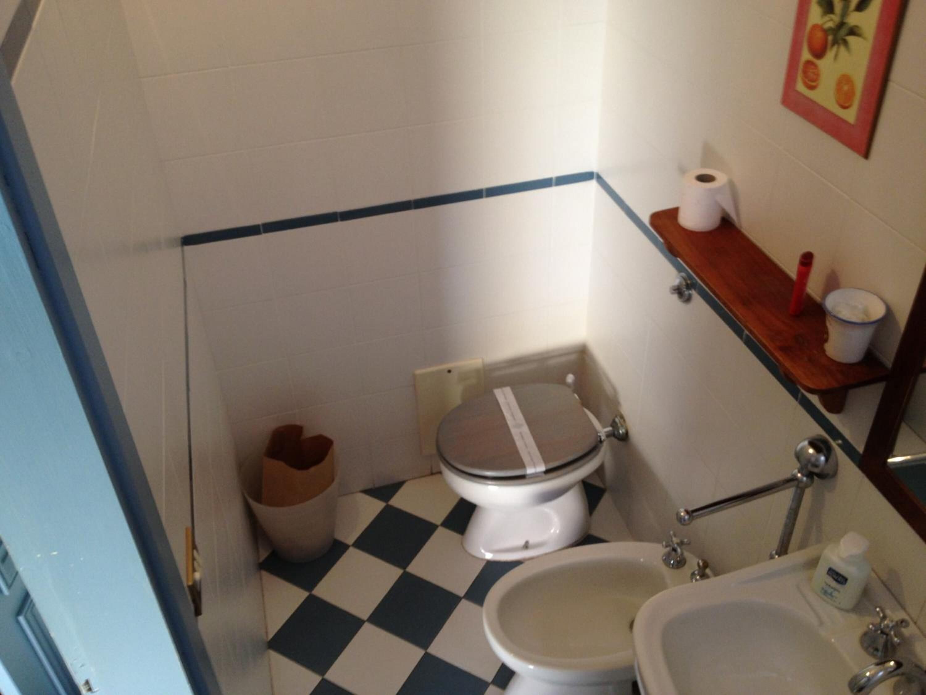 A small bathroom with a toilet, bidet, and sink. There is a trash bin and a wooden shelf with a roll of toilet paper and a red object.