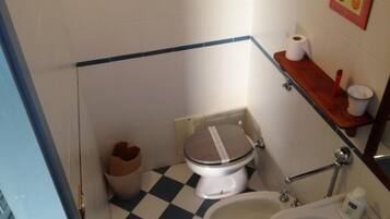 A small bathroom with a toilet, bidet, and sink. There is a trash bin and a wooden shelf with a roll of toilet paper and a red object.
