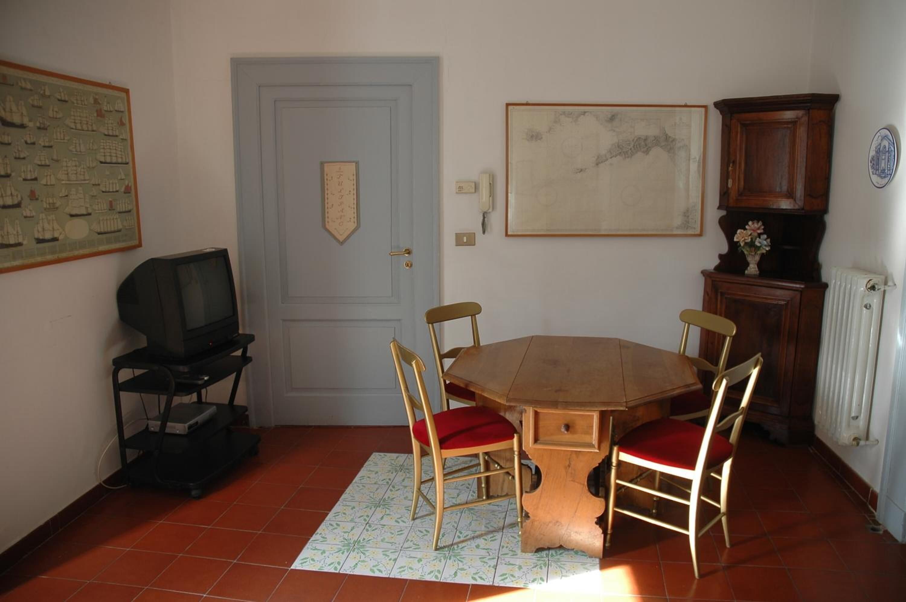 Zona pranzo con tavolo in legno e quattro sedie, televisore su un mobile, porta con stemma e quadro appeso al muro.
