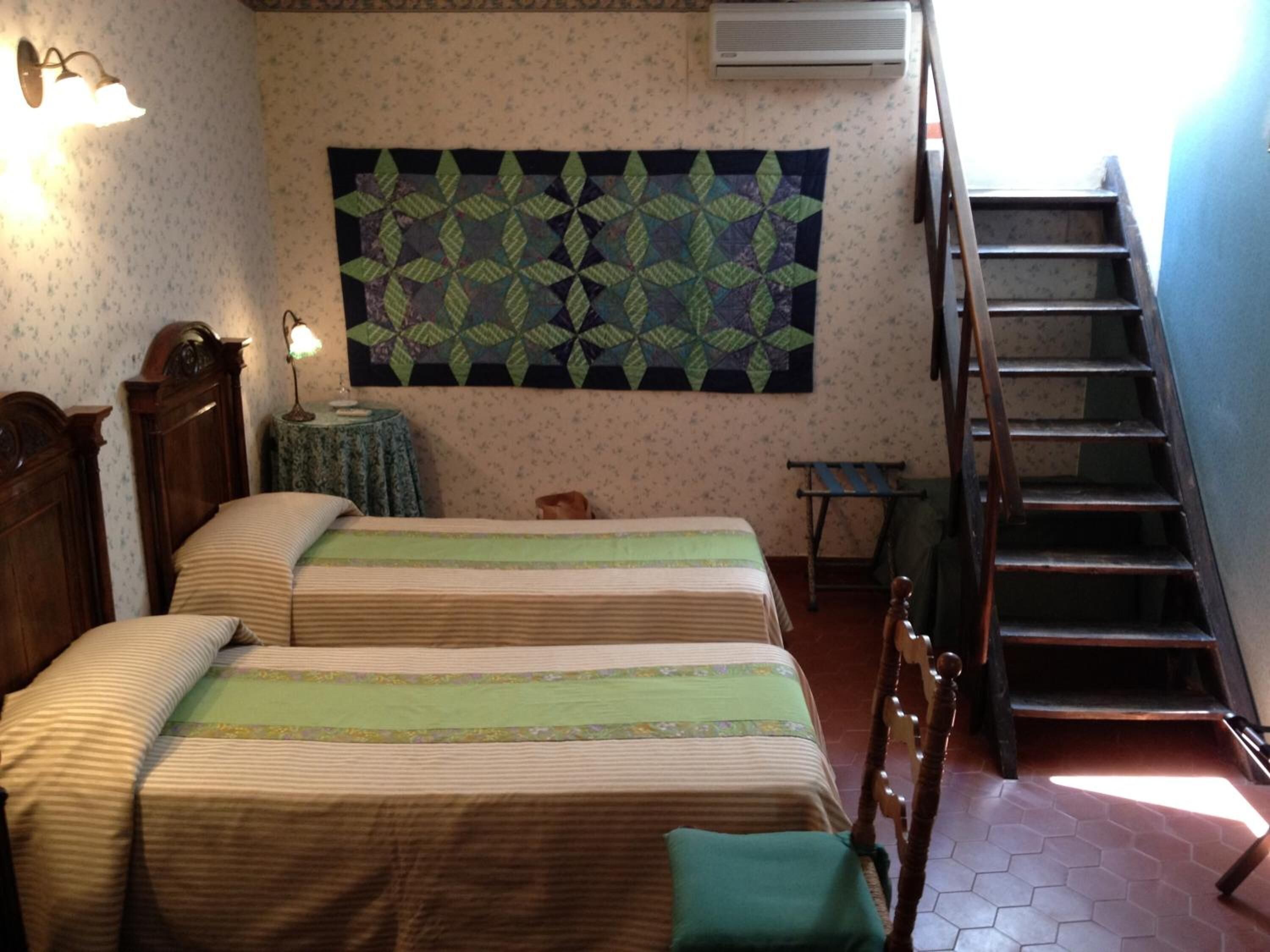 A bedroom with two beds, a wall hanging, a small table, a lamp, and a staircase.