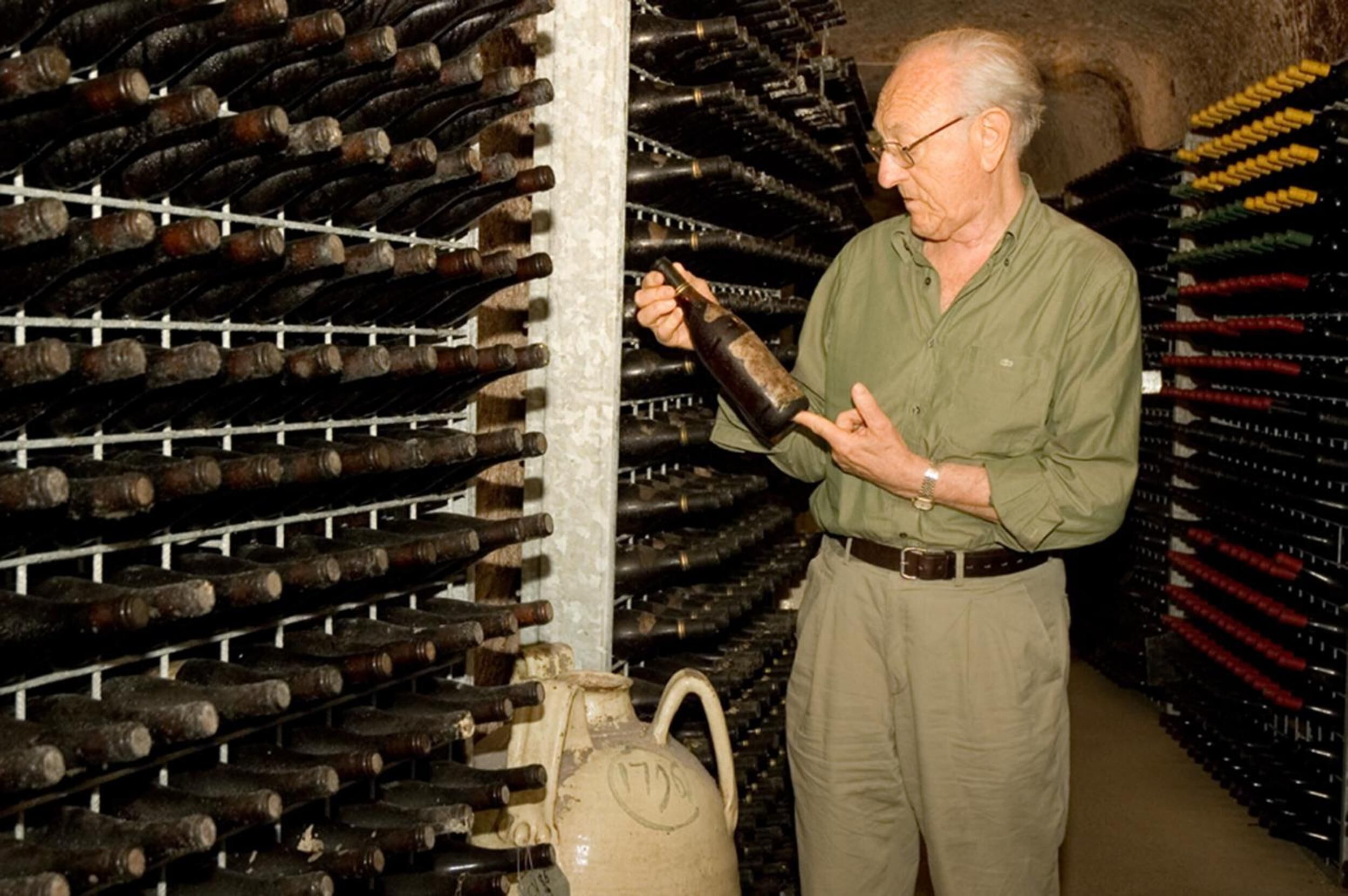 A man inspecting a bottle in a wine cellar with rows of wine bottles and a large ceramic jug.