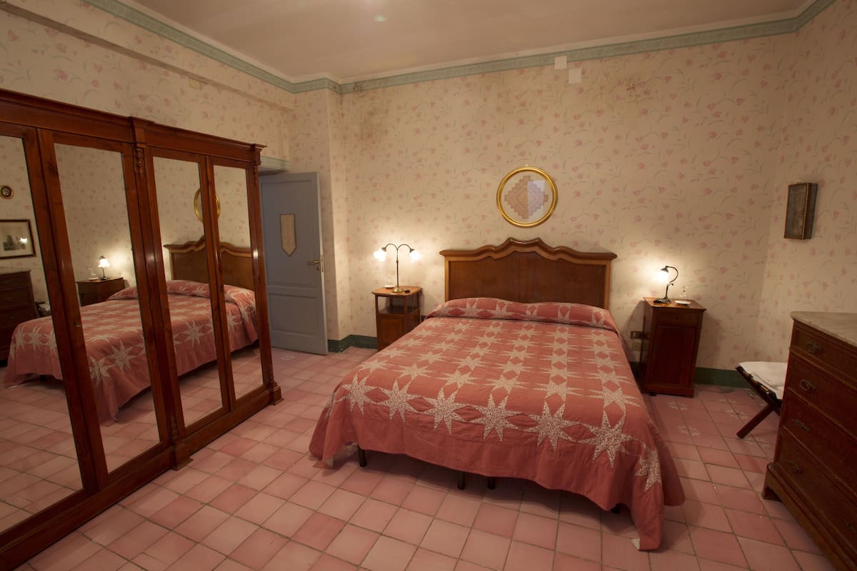 Une chambre à coucher avec un grand miroir, un lit recouvert d’une couvre-lit rose, une tête de lit en bois et deux tables de chevet avec des lampes.