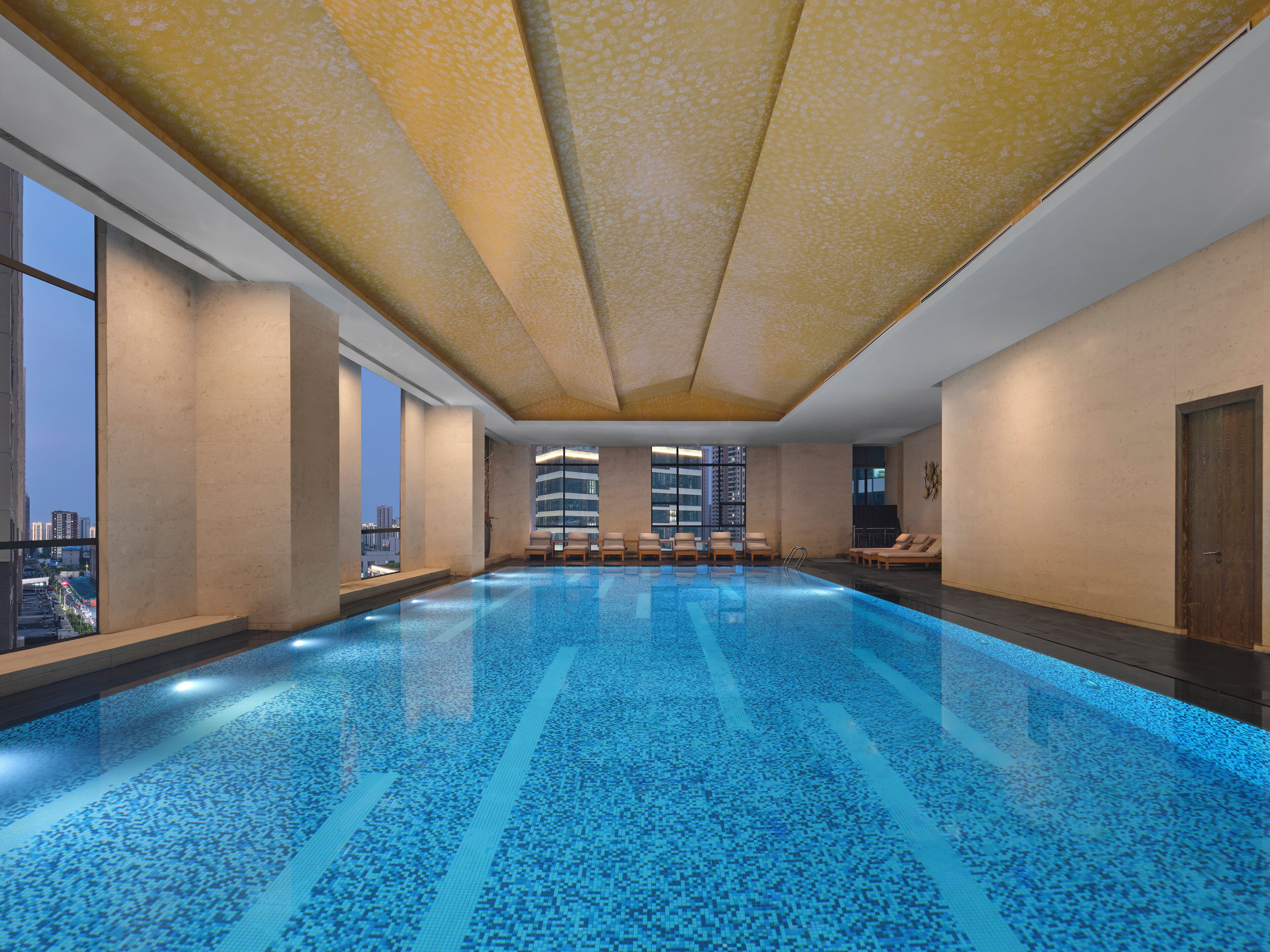 An indoor swimming pool with clear blue tiles, large windows, and a view of a cityscape.
