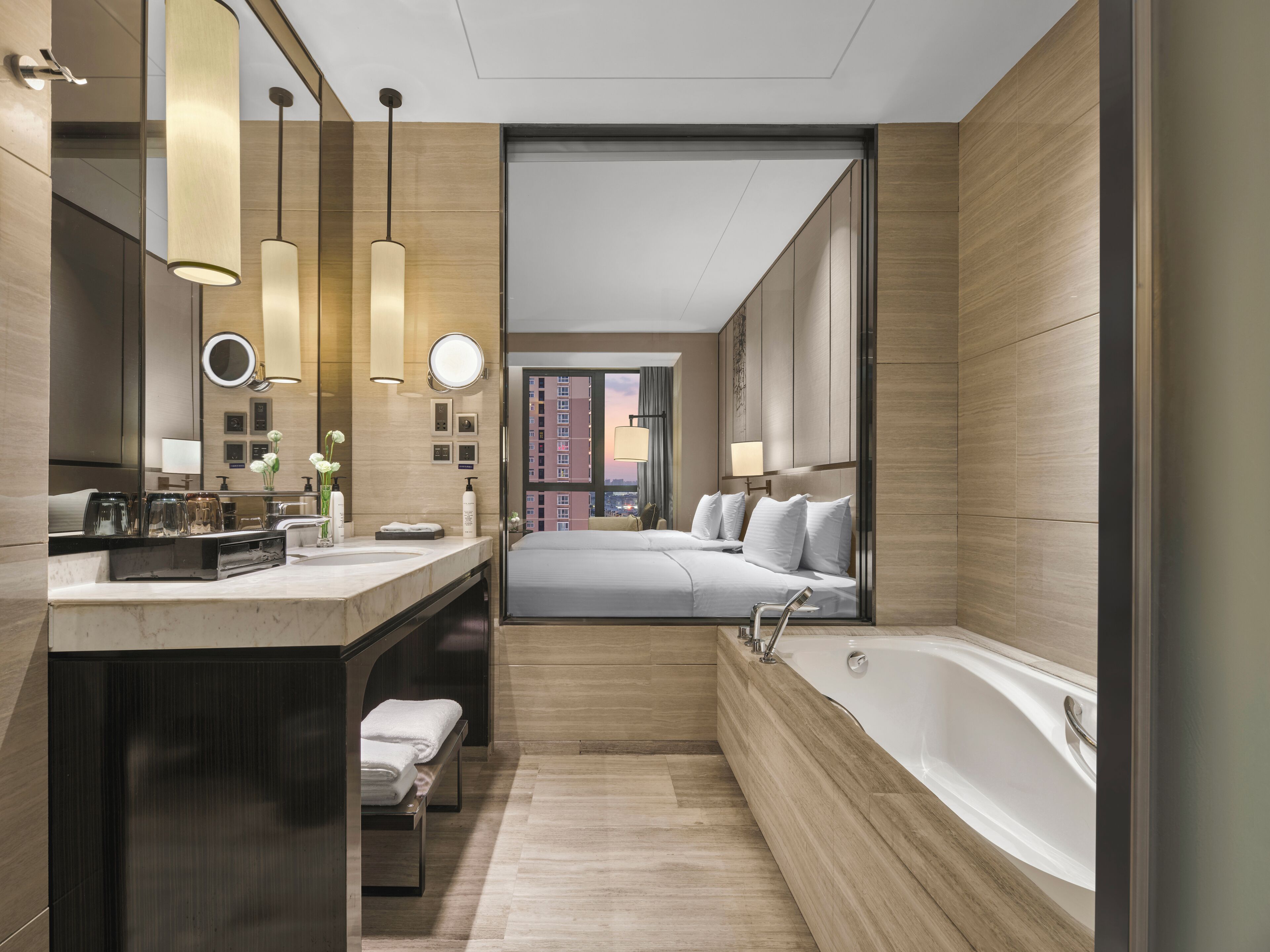 A modern hotel bathroom with a large bathtub, a vanity with a mirror, and a view of the cityscape through a large window.