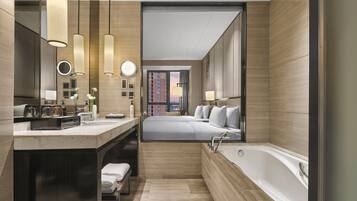 A modern hotel bathroom with a large bathtub, a vanity with a mirror, and a view of the cityscape through a large window.