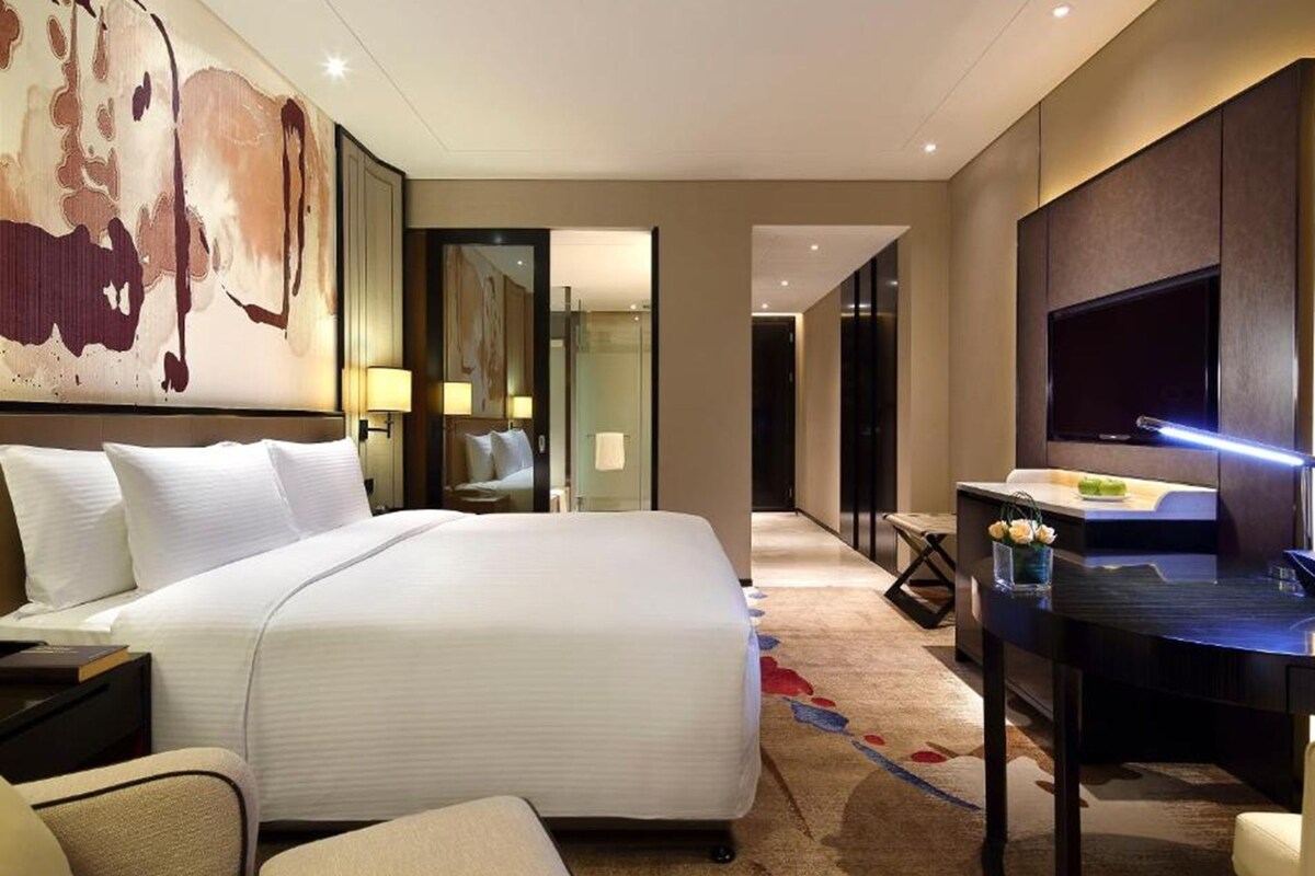 A hotel room with a large bed, a desk with a TV, and a painting on the wall.