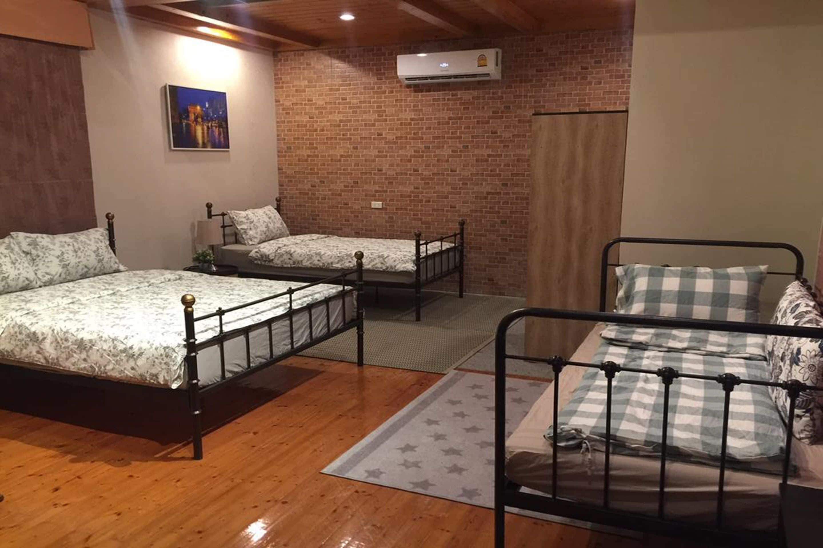 Quarto com três camas de metal, piso de madeira, parede de tijolos e ar-condicionado.