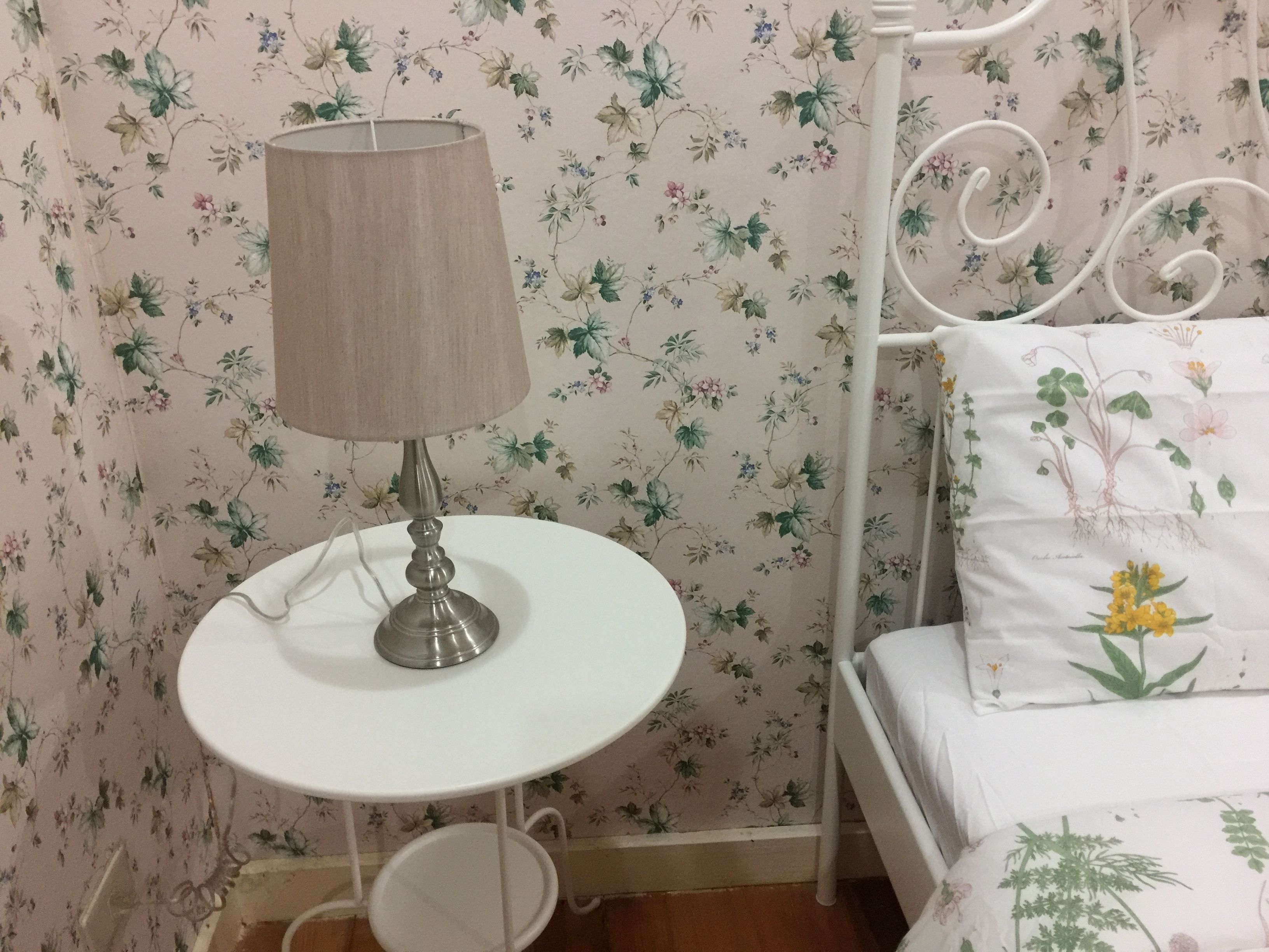 Una mesa redonda pequeña con una lámpara, una cama con ropa de cama floral y un fondo de papel tapiz con motivos florales.
