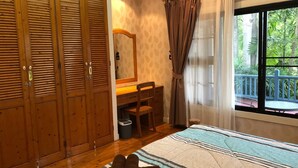 Ein Schlafzimmer mit Holzschrank, Schreibtisch mit Stuhl, Bett mit gestreifter Bettdecke und Fenster mit Blick ins Grüne.