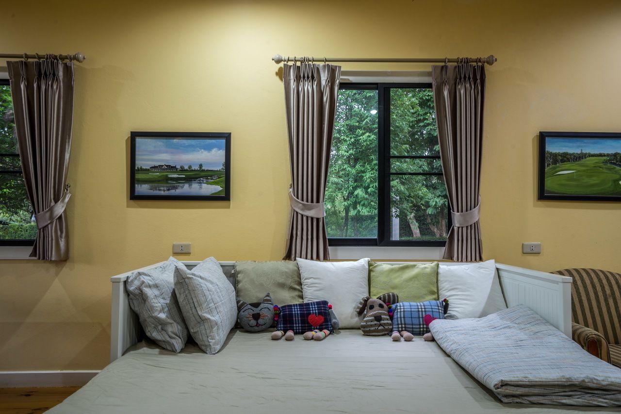 A bedroom with a bed, pillows, and stuffed animals. There are two windows with curtains, and two framed pictures on the wall.