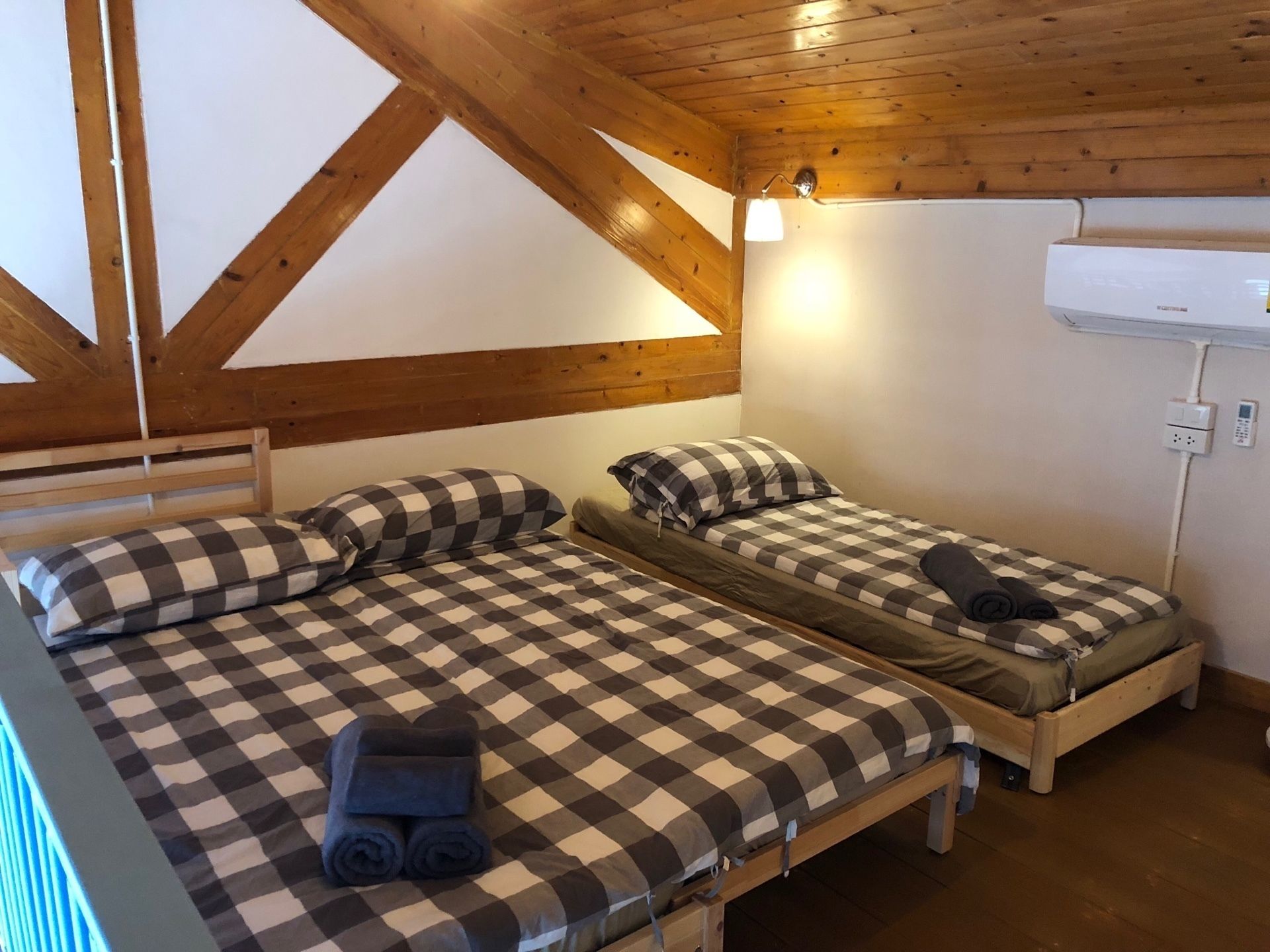 A room with two beds, each with a checkered bedspread and pillows. There is a wall-mounted air conditioning unit and a light fixture above the beds.