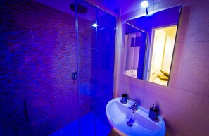 Un baño moderno con cabina de ducha de vidrio, un lavabo blanco y un espejo que refleja una habitación con cama.