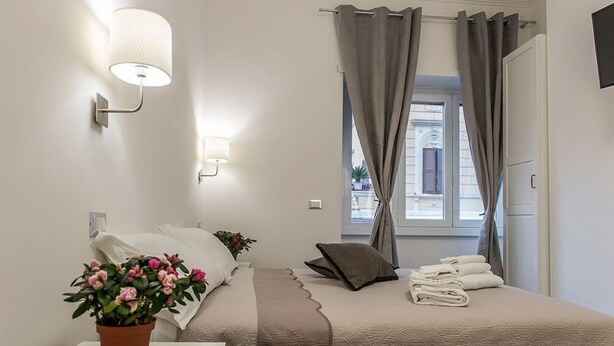 A neatly arranged bedroom with a bed, a bedside table with a potted plant, a lamp, and a window with curtains. - Mr. Charlie Brown (Rome)