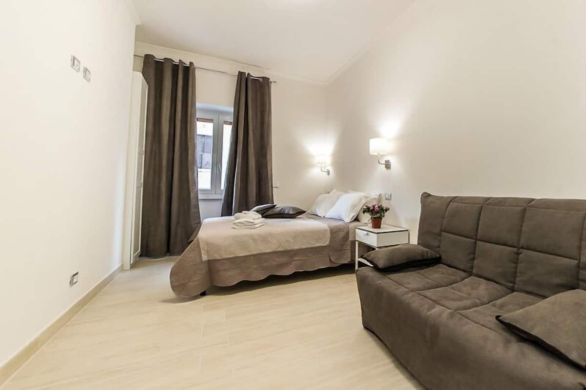 A small, modern bedroom with a bed, a sofa, a nightstand, and a window with curtains.