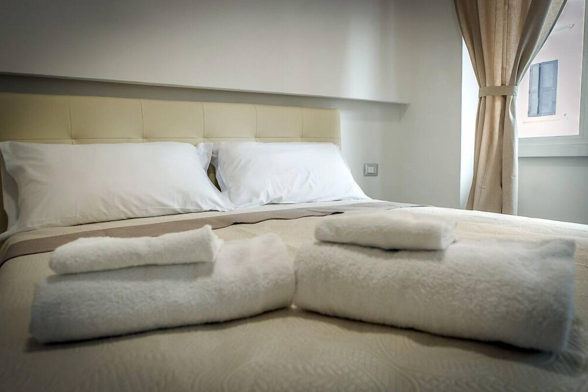 A neatly made bed with white linens and towels, a beige headboard, and a window with curtains.