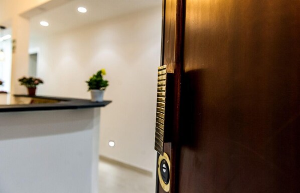 Una puerta de madera con manija de bronce y cerradura, que da acceso a una zona de recepción con mostrador y plantas en macetas.